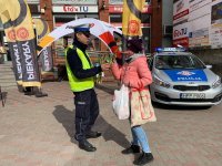 Zdjęcie kolorowe. Na zdjęciu widoczny policjant wręczający tulipana kobiecie. W otoczeniu widoczne placówki handlowe. Zdjęcie zrobione na chodniku przy ulicy Wyszyńskiego.
